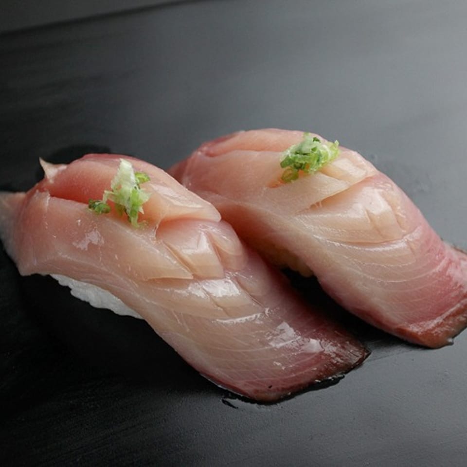 Two pieces of yellowtail nigiri sushi with light pink fish and green garnish on a dark surface.