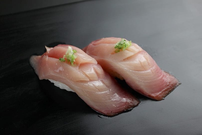 Two pieces of yellowtail nigiri sushi with light pink fish and green garnish on a dark surface.