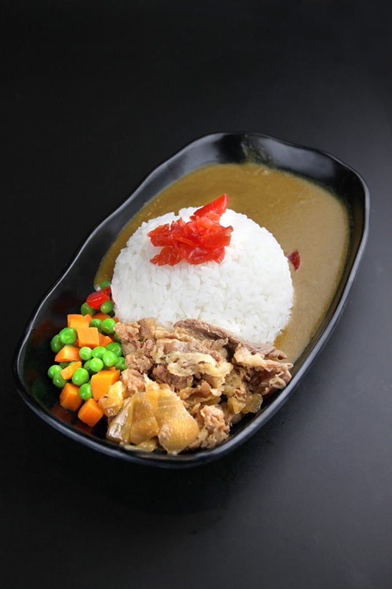 Japanese curry with rice, beef, pickled ginger, peas, and carrots in a black dish.
