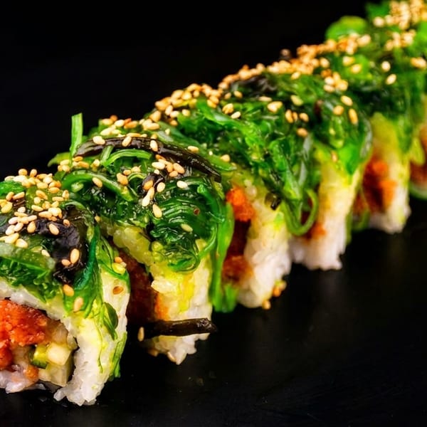 Sushi roll with green seaweed salad and sesame seeds on a dark background.