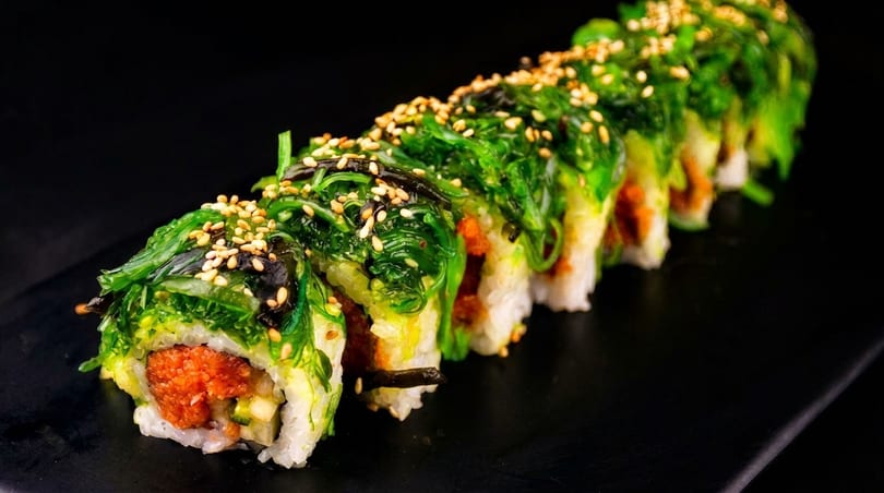 Sushi roll with green seaweed salad and sesame seeds on top, filled with orange spicy tuna.