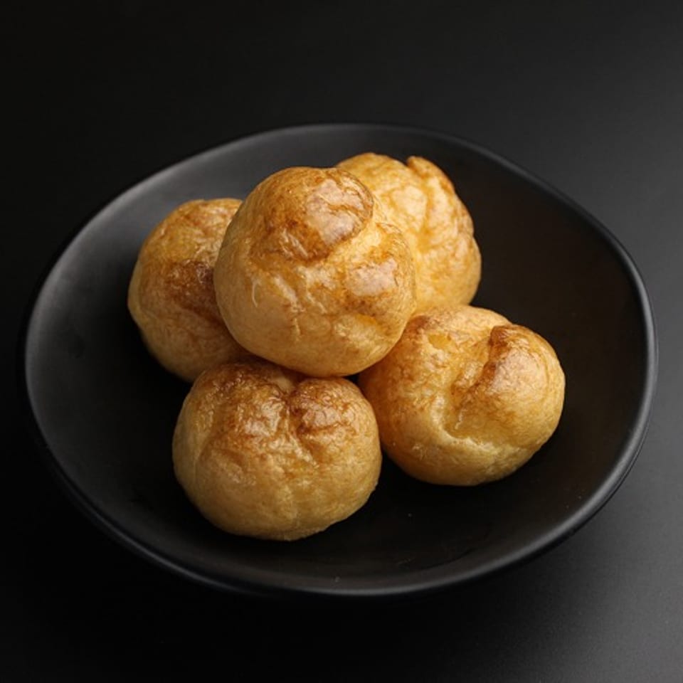 Five golden-brown gougères, or cheese puffs, are arranged on a black plate against a dark background.