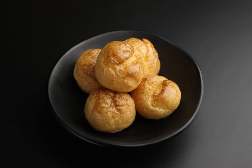 Five golden-brown gougères, or cheese puffs, are arranged on a black plate against a dark background.