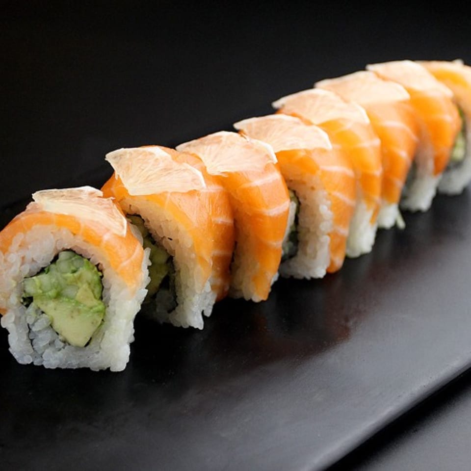 A row of salmon and avocado sushi rolls with thin lemon slices on a dark plate.