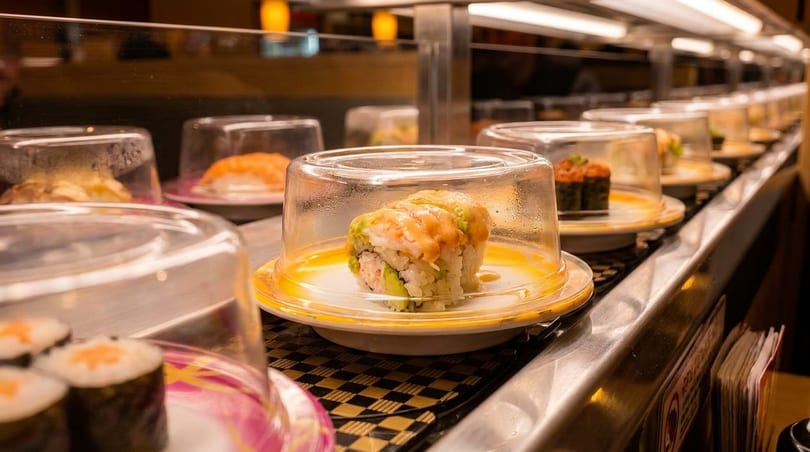 Sushi plates with clear covers move along a conveyor belt in a restaurant.
