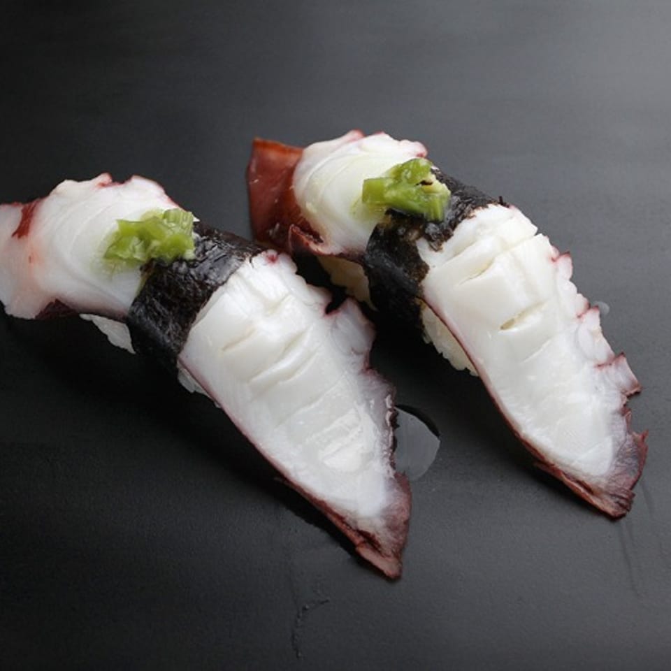 Two pieces of octopus nigiri sushi with wasabi and nori on a dark plate.