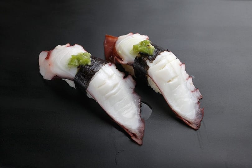 Two pieces of octopus nigiri sushi with wasabi and nori on a dark plate.