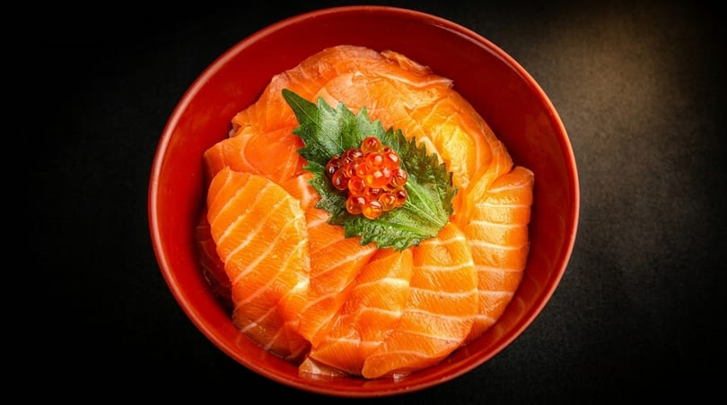 Salmon sashimi slices arranged in a red bowl, topped with green shiso leaf and salmon roe.