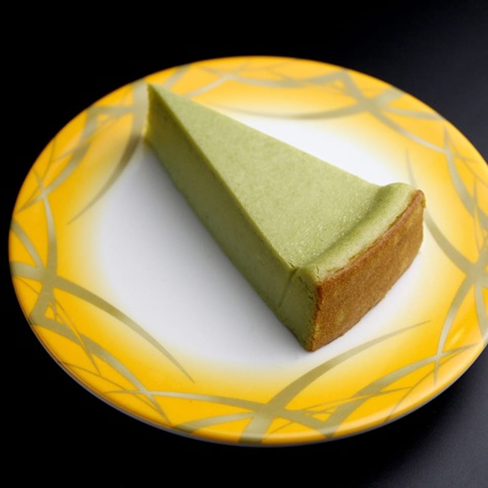A slice of green matcha cheesecake with a brown crust on a yellow and white patterned plate.
