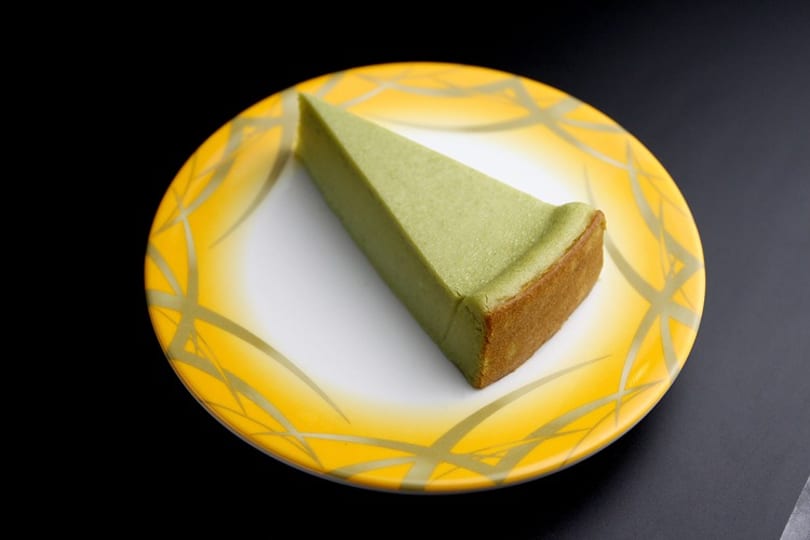 A slice of green matcha cheesecake with a brown crust on a yellow and white patterned plate.