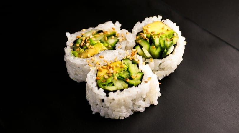 Three sushi rolls with avocado, cucumber, and sesame seeds on a dark surface.