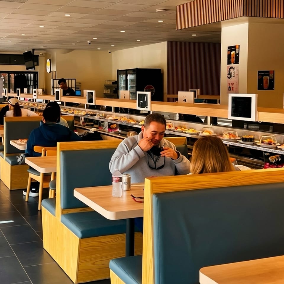 Diners seated at booths in a modern sushi restaurant with a conveyor belt displaying various dishes.